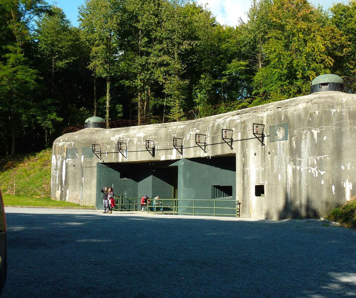 A large, weathered concrete bunker with green steel doors is built into a hillside surrounded by trees, with a few visitors standing outside.