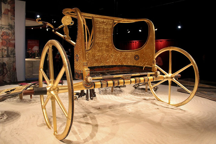 Gold-adorned ancient Egyptian chariot on display, featuring engraved symbols, decorative elements, and spoked wheels.