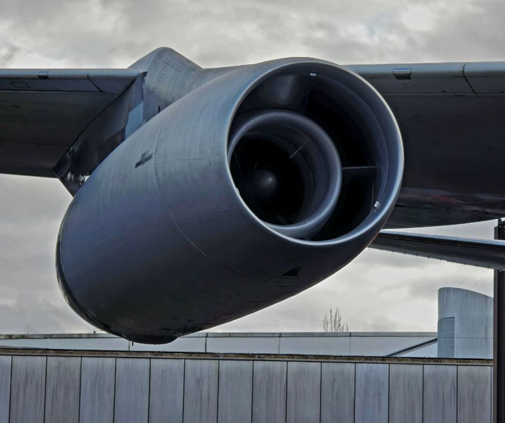 A close-up of a jet engine mounted under the wing of an aircraft. The turbine fan is visible inside the engine, with a cloudy sky and part of the fuselage in the background.