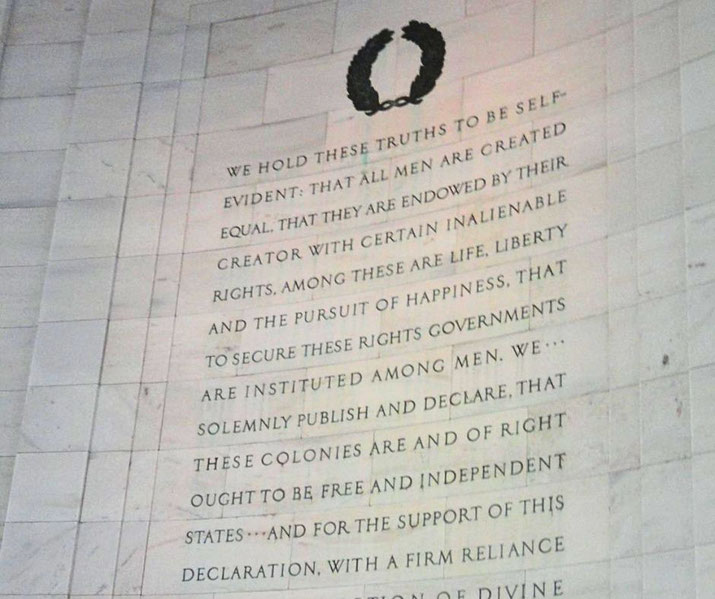 Engraved wall text from the Declaration of Independence with a laurel wreath above, emphasizing liberty and equality in a historic monument setting.