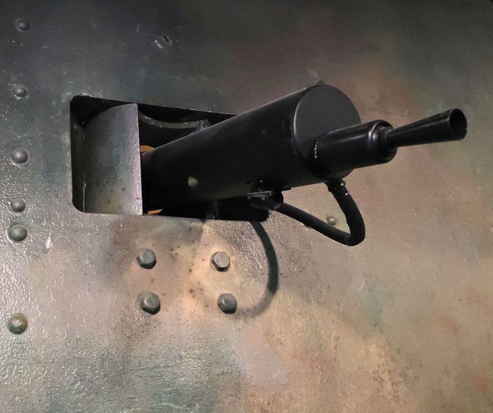 A machine gun protrudes from an armored vehicle or bunker wall, surrounded by rivets and painted metal plating.