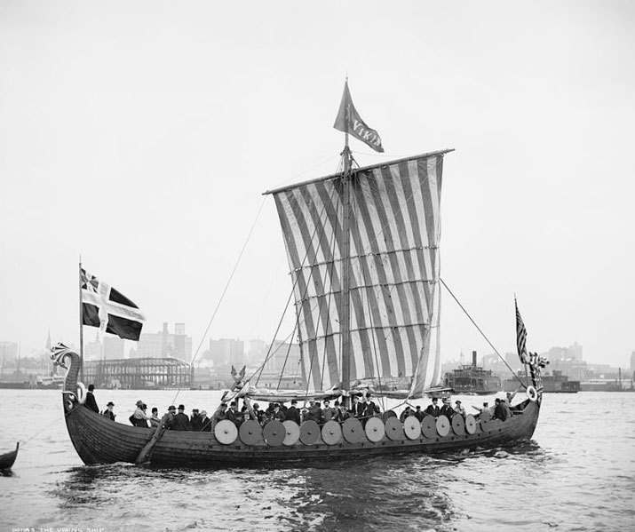 A recreated Viking longship sailing on a river with a striped sail, round shields along the hull, and a dragon-headed prow.