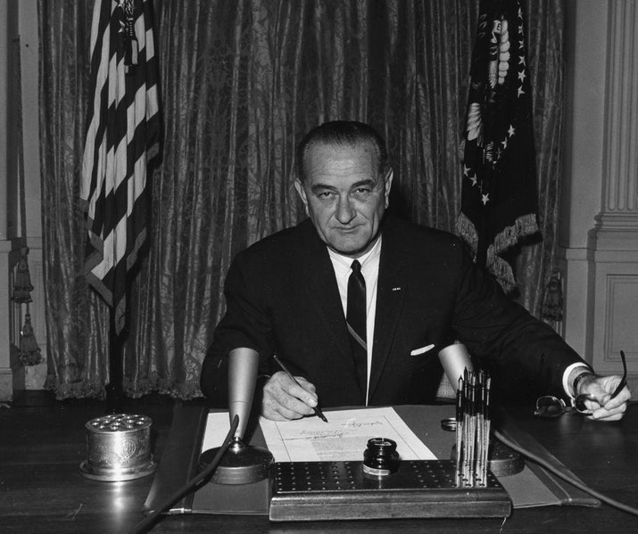 A U.S. president sits at a desk signing a document, flanked by American and presidential flags in the background.