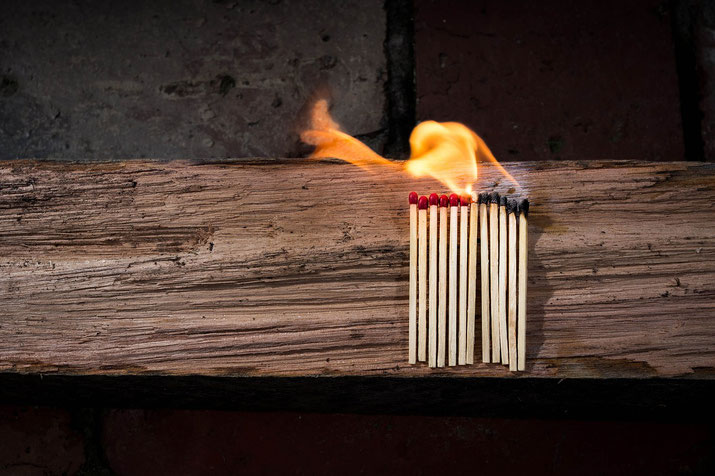 A row of matches is lined up on a wooden surface, with one side burned and the fire spreading towards the unlit matches.