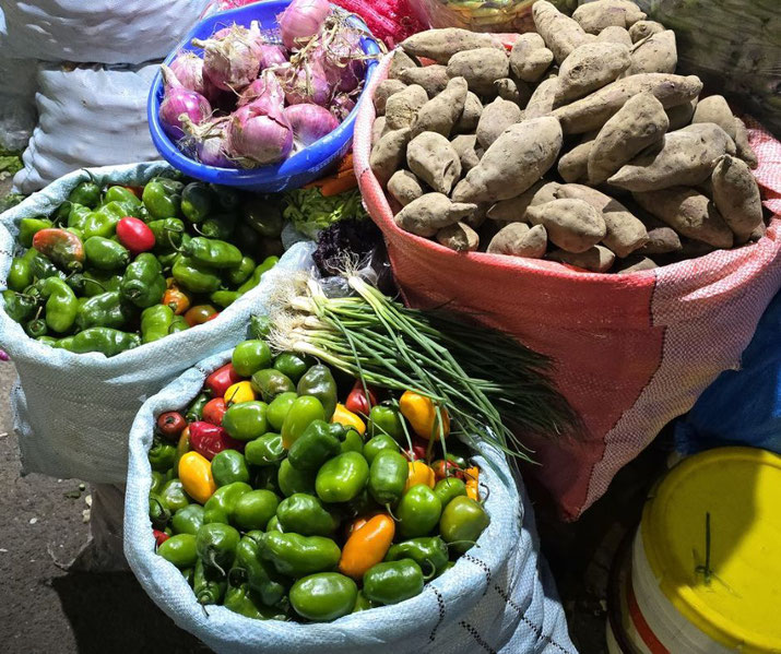 A market stall with sacks of fresh produce, including sweet potatoes, green and colorful chili peppers, spring onions, and a bowl of red onions. The setting appears to be outdoors.