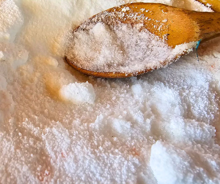 A wooden spoon scoops coarse white salt from a pile, showing grainy texture and natural crystal clumps.