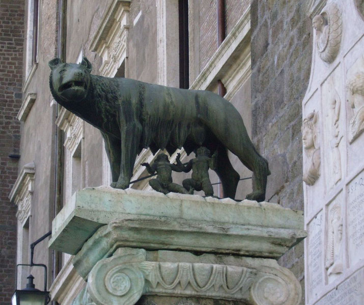 The Capitoline Wolf, a bronze statue of a she-wolf suckling the twins Romulus and Remus, symbolic of Rome's foundation myth, mounted on a decorated pedestal near a stone relief.