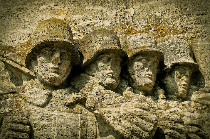 Stone relief sculpture of four soldiers wearing helmets, carved into a wall, with solemn expressions and weathered textures.