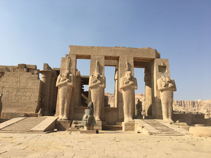 Stone statues of pharaohs stand in front of an ancient Egyptian temple with hieroglyphs and columns under a clear sky.