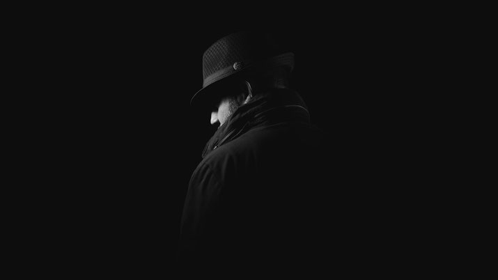 A man in a fedora and coat faces away in low-key lighting, creating a moody, mysterious silhouette against a dark background.