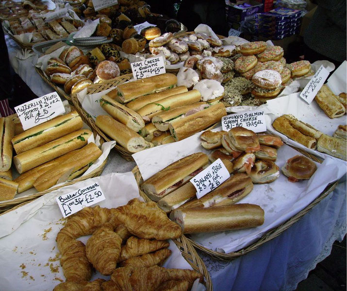 A variety of freshly baked goods on display at a market stall, including sandwiches, bagels, croissants, and pastries, with price labels indicating different items like smoked salmon and ham cheese.