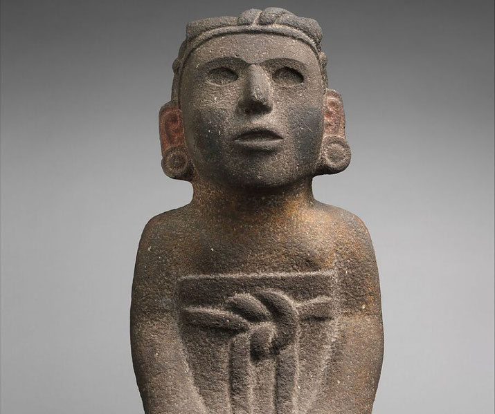 An ancient stone statue features a human-like figure with a solemn expression, large ears, and intricate carvings on its body.