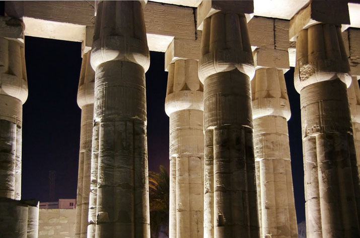 Massive ancient Egyptian columns illuminated at night, featuring lotus-shaped capitals and engraved hieroglyphs.