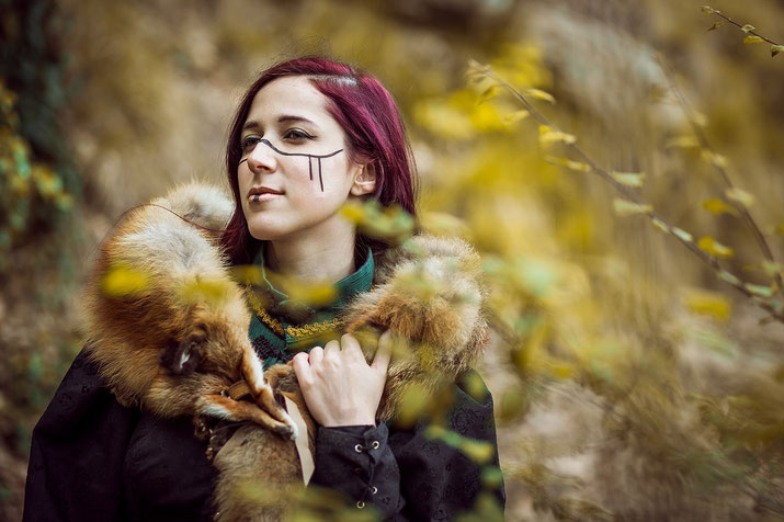 A person with red hair and face paint wearing a fur stole, set in a natural, forested environment. They exude a medieval or Viking-inspired aesthetic, with a serene, confident expression.