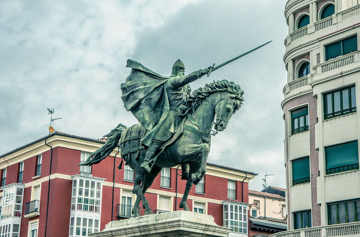 Equestrian statue of a medieval knight with raised sword, surrounded by colorful and modern buildings.