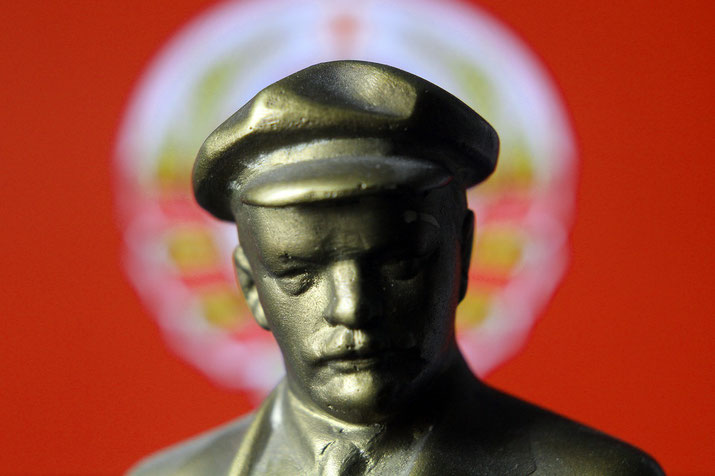 Close-up of a small metallic Lenin bust wearing a cap, set against a blurred red Soviet emblem background.