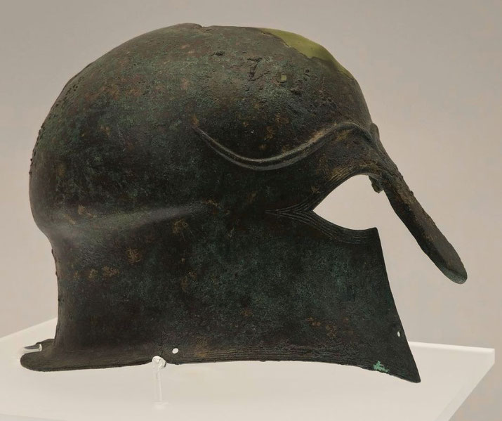 Ancient bronze helmet with a prominent nose guard and eye openings, showing a worn, green patina and smooth rounded shape.