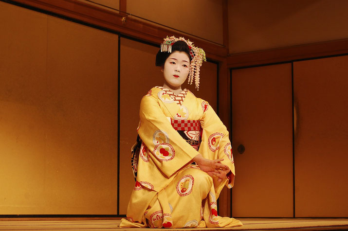 A Maiko in a vibrant yellow kimono with intricate patterns performs a traditional dance. She wears elaborate hair ornaments, white makeup, and a red obi.