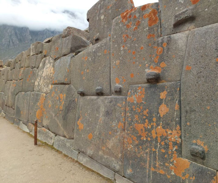 An ancient Inca stone wall at Ollantaytambo, Peru, featuring precisely cut stones with protruding knobs and patches of orange lichen.