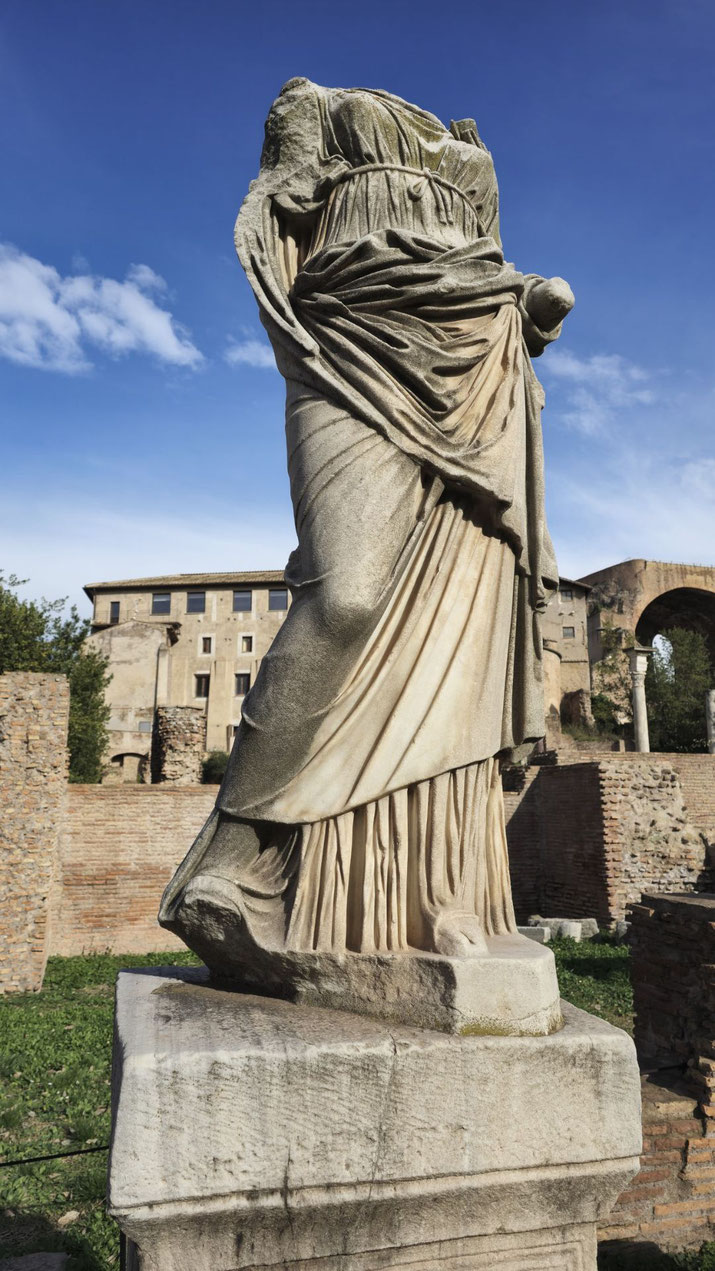 Headless ancient statue of a robed figure with one arm missing, set on a stone pedestal amid Roman ruins under a clear blue sky.