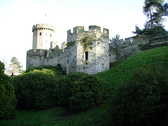A view of stone castle walls and a round tower surrounded by greenery, with lush grass and trees enhancing the medieval fortification's serene yet imposing atmosphere.