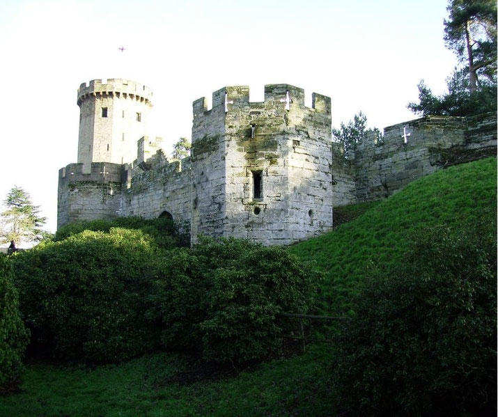 A view of stone castle walls and a round tower surrounded by greenery, with lush grass and trees enhancing the medieval fortification's serene yet imposing atmosphere.