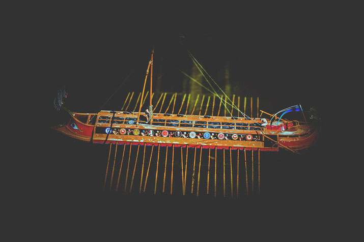 Model of an ancient warship with colorful shields and multiple oars, lit dramatically against a dark background.