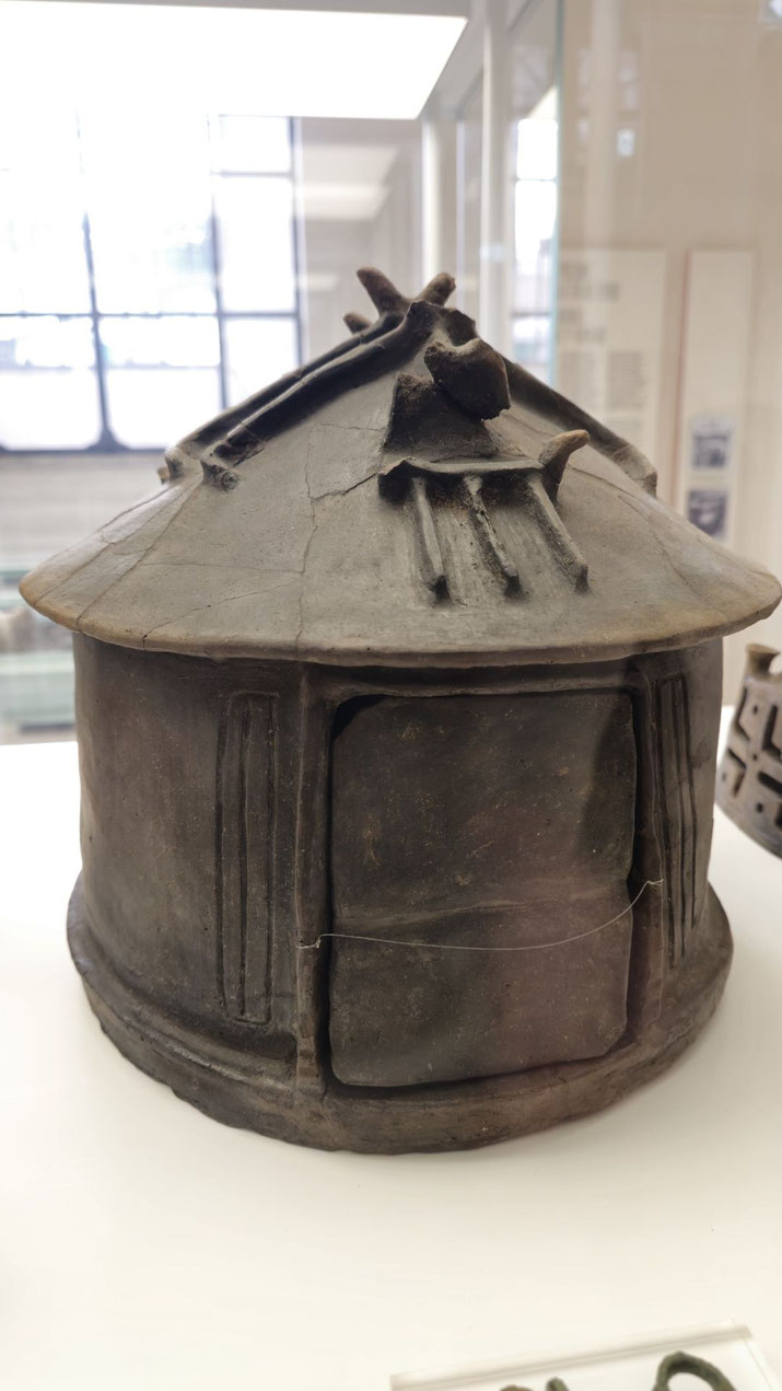 Ancient clay model of a round hut with a conical roof, central door, and stylized architectural details, possibly representing an early Italic or Etruscan dwelling.