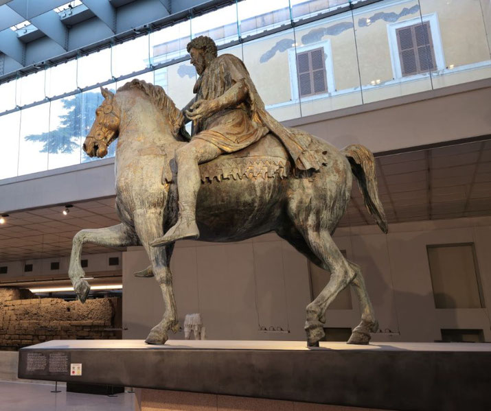 Large bronze equestrian statue of a bearded man in a toga, with one arm extended, seated on a trotting horse.