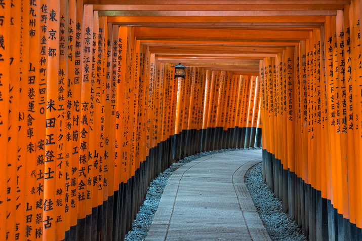 A winding stone path passes through rows of vivid orange torii gates inscribed with black Japanese characters at a famous shrine.
