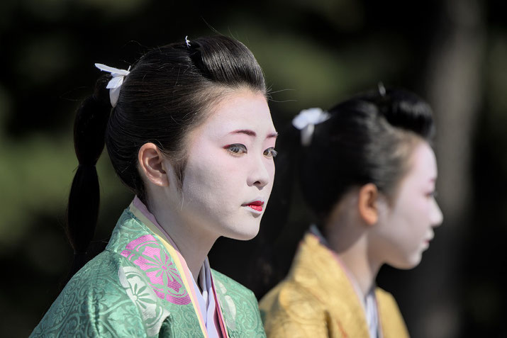 Two women dressed in traditional Japanese attire wear elegant kimonos with intricate patterns. Their faces are painted white with red and black accents, and their hair is styled neatly with decorative accessories.