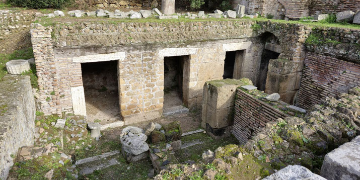 Ancient stone tombs with three arched doorways, surrounded by moss-covered ruins and scattered stone fragments.