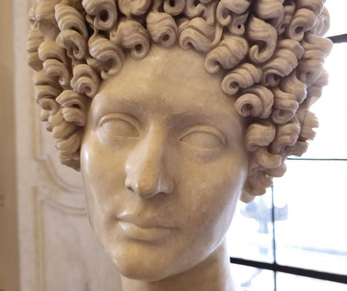 Marble bust of a woman with a calm expression and an elaborate hairstyle of tight, voluminous curls, showcasing Roman sculptural detail.