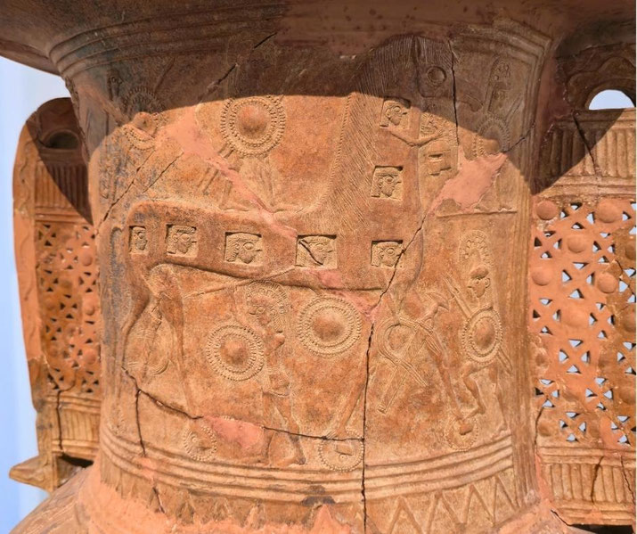 Ancient terracotta relief on the Mykonos Vase depicting armed warriors, shields, and the Trojan Horse with carved figures emerging from its body.
