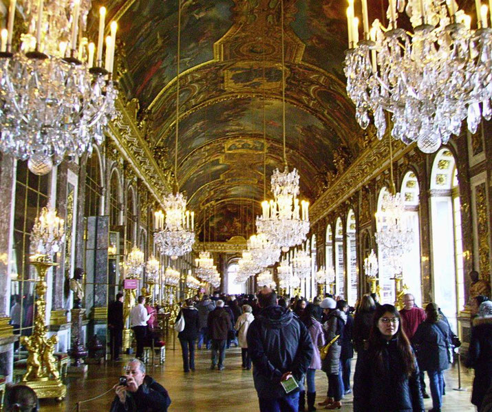 Visitors walk through a grand hallway with arched ceilings, chandeliers, mirrors, and gilded decor.