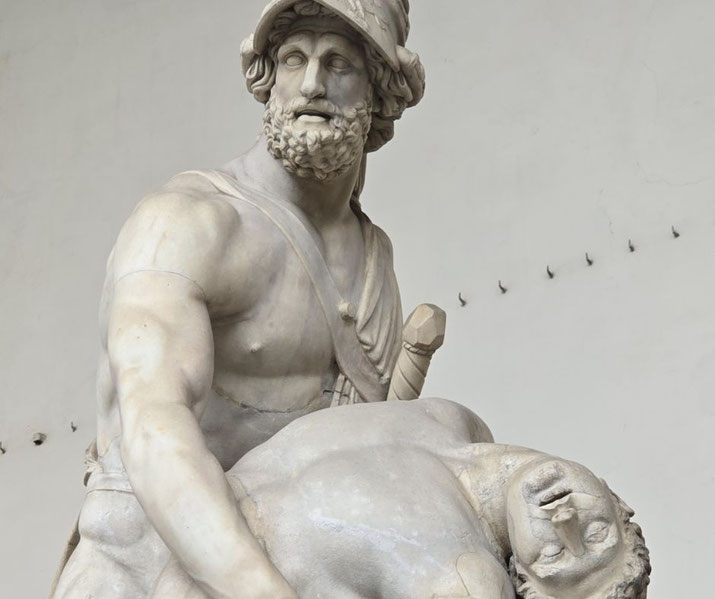 Marble statue of a muscular warrior in a helmet holding a lifeless male figure, with a sword sheathed at his side.