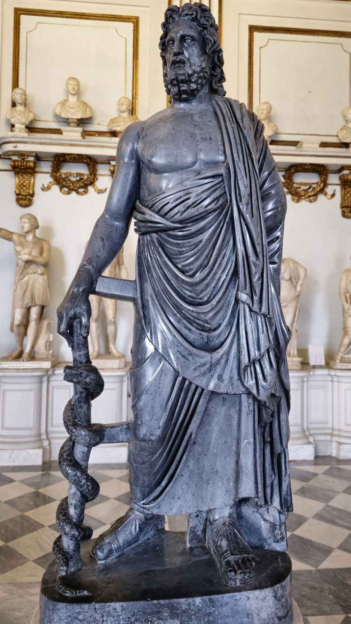 Dark stone statue of a bearded man in draped robes holding a staff with a coiled serpent, symbolizing healing or medicine, likely representing the god Asclepius.