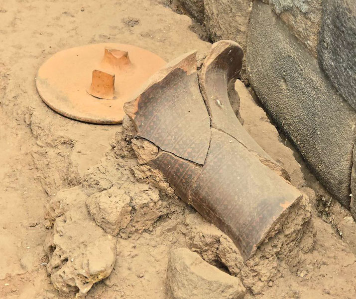 Broken ancient pottery with geometric patterns lies partially buried in soil next to a round ceramic lid and stone wall, likely part of an archaeological dig.