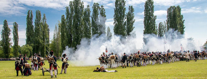 Historical reenactors in Napoleonic-era uniforms engage in a mock battle on a grassy field with smoke rising and trees in the background.