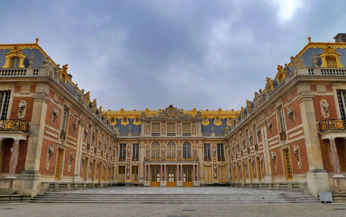 The grand facade of the Palace of Versailles, featuring gold-accented rooftops, ornate windows, and symmetrical classical architecture under a cloudy sky.