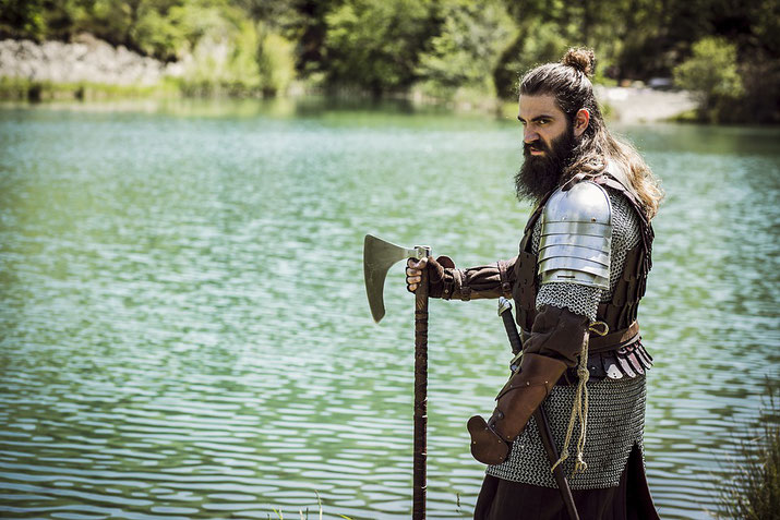 Man dressed as a Viking warrior stands by a lake holding a battle axe, wearing chainmail and armor with a stern expression.