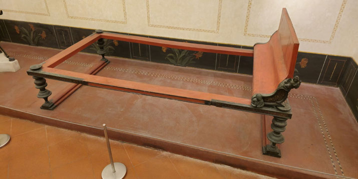 Ancient Roman wooden bed frame with ornate bronze legs and backrest, placed in a room with decorative floral wall and floor patterns.