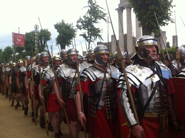 A reenactment of Roman legionaries marching in formation, wearing lorica segmentata armor and red tunics, carrying spears and shields, with a banner reading "LEG XI" visible in the background.