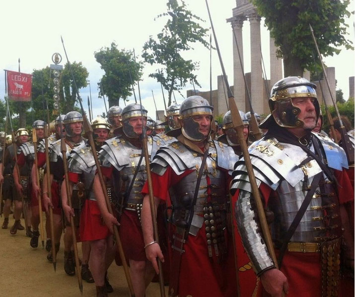 A reenactment of Roman legionaries marching in formation, wearing lorica segmentata armor and red tunics, carrying spears and shields, with a banner reading "LEG XI" visible in the background.