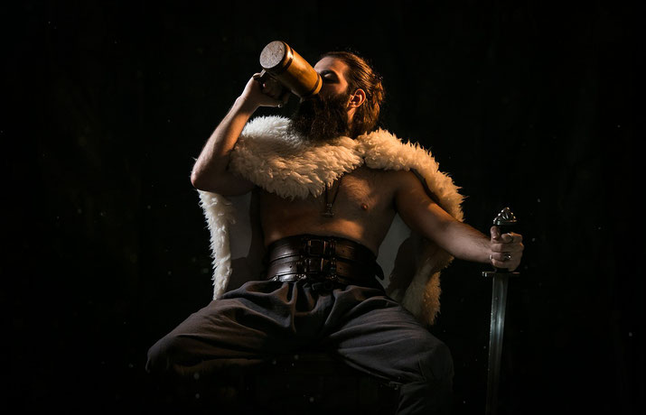 A bearded warrior sits confidently, draped in a fur cloak, drinking from a wooden tankard.