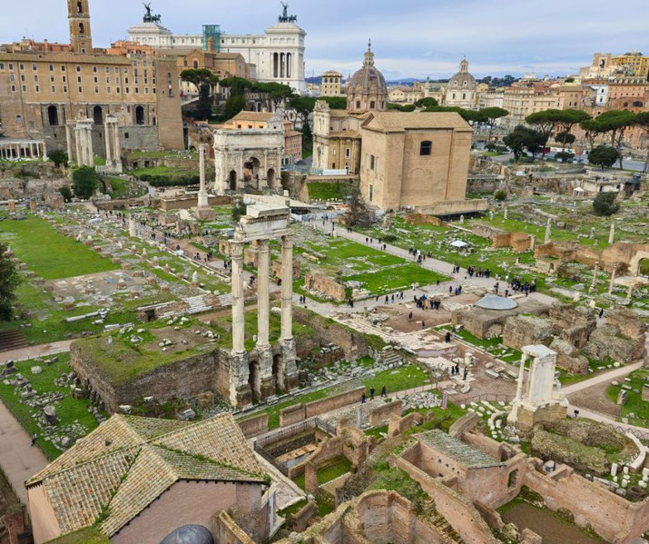 Ancient Roman Forum ruins with scattered columns, arches, and temples surrounded by modern buildings under a cloudy sky.