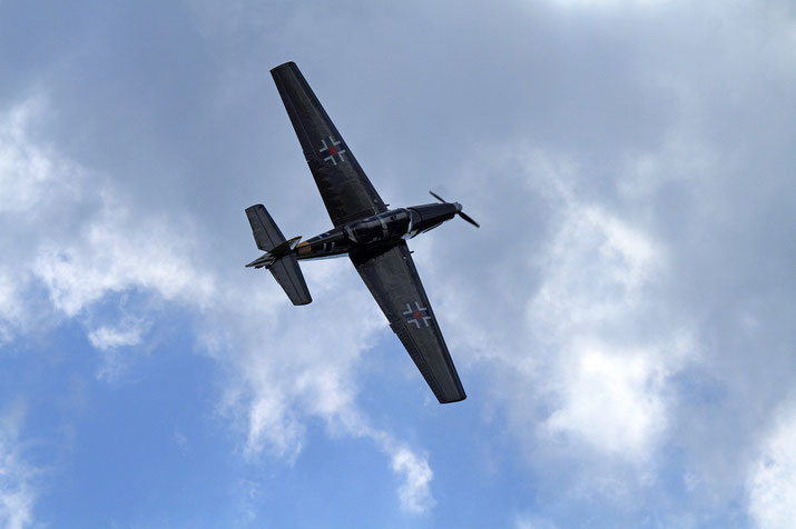 A dark military-style aircraft with cross insignias flies overhead against a partly cloudy sky.