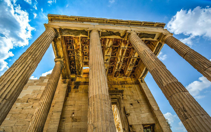 Low-angle view of tall ancient Greek columns and a weathered coffered ceiling set against a bright blue sky with clouds.