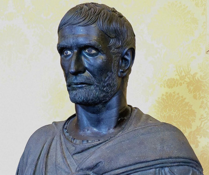 Dark bronze bust of a bearded man with deep-set eyes and short hair, wearing a draped garment, set against a light patterned background.