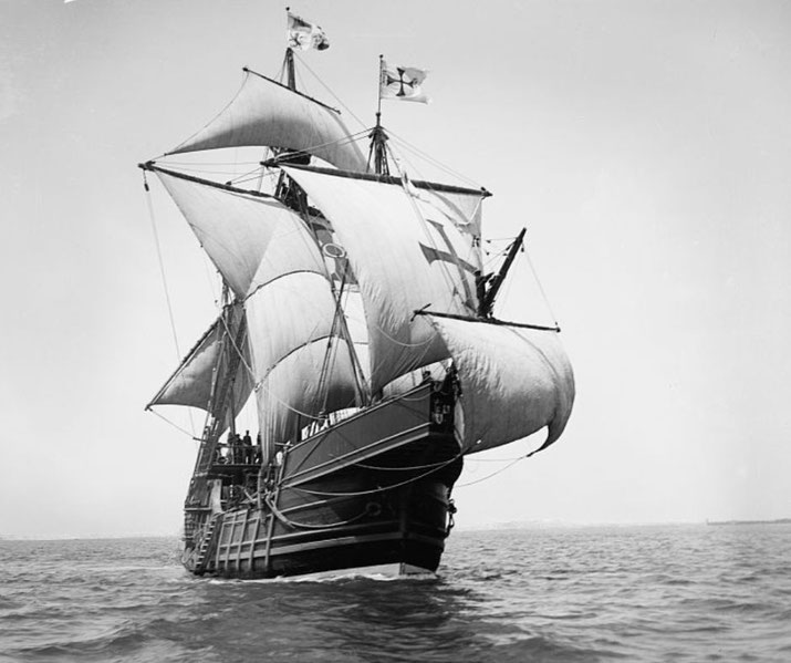 A replica of a Spanish caravel sailing with full sails. The ship, reminiscent of Columbus' Santa María, features a high stern, square-rigged masts, and fluttering flags.